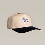 The Shop Hat | Pointer by Ballad of the Bird Dog is a beige and black embroidered hat featuring a blue pointing bird dog on the front, ideal for outdoor adventures, shown against a plain light background.