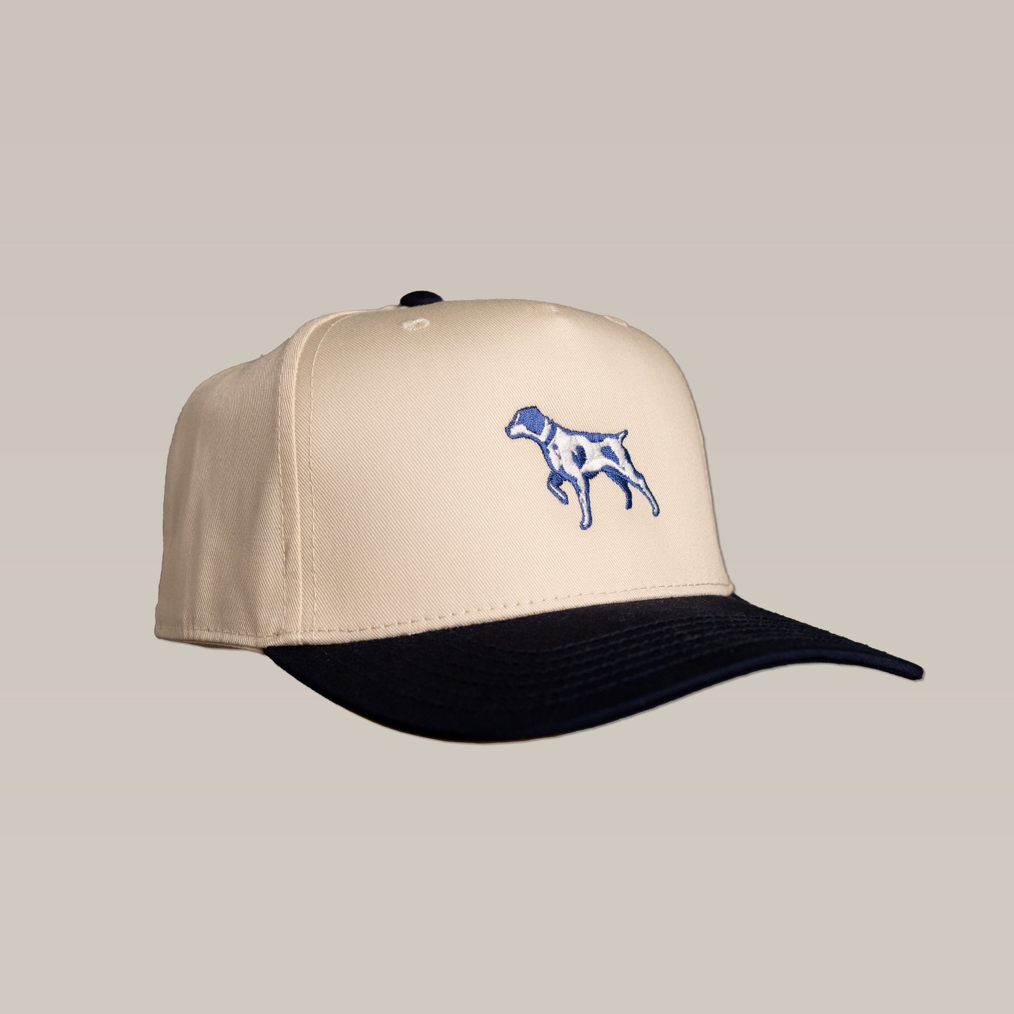 The Shop Hat | Pointer by Ballad of the Bird Dog features a beige crown, navy brim, and blue embroidered dog—ideal for outdoor adventures. Displayed against a simple background.