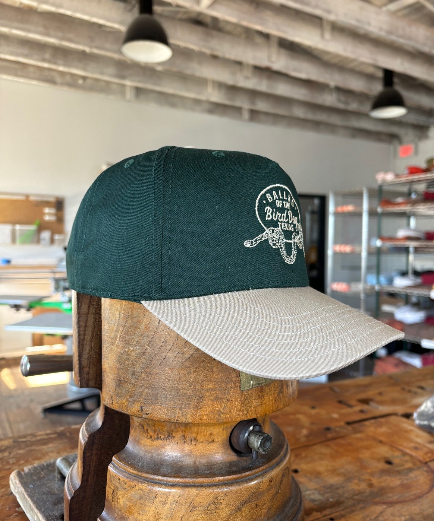 The Ballad of the Bird Dog Shop Hat featuring a Rattlesnake Oval Logo from the desert collection is displayed on a wooden form, highlighting the handcrafted quality Brenham, TX is known for.