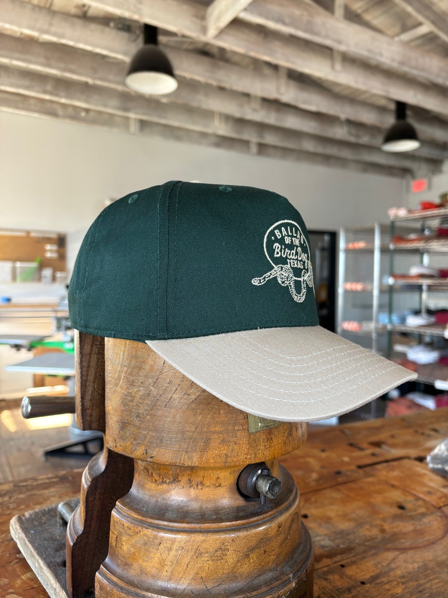 The Ballad of the Bird Dog Shop Hat featuring a Rattlesnake Oval Logo from the desert collection is displayed on a wooden form, highlighting the handcrafted quality Brenham, TX is known for.