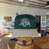 The Shop Hat | Rattlesnake Oval Logo by Ballad of The Bird Dog from the Desert Collection, in green and beige with a rattlesnake graphic and "Dallas by the Bird Dog, Texas," rests on a wooden stand in a workshop.