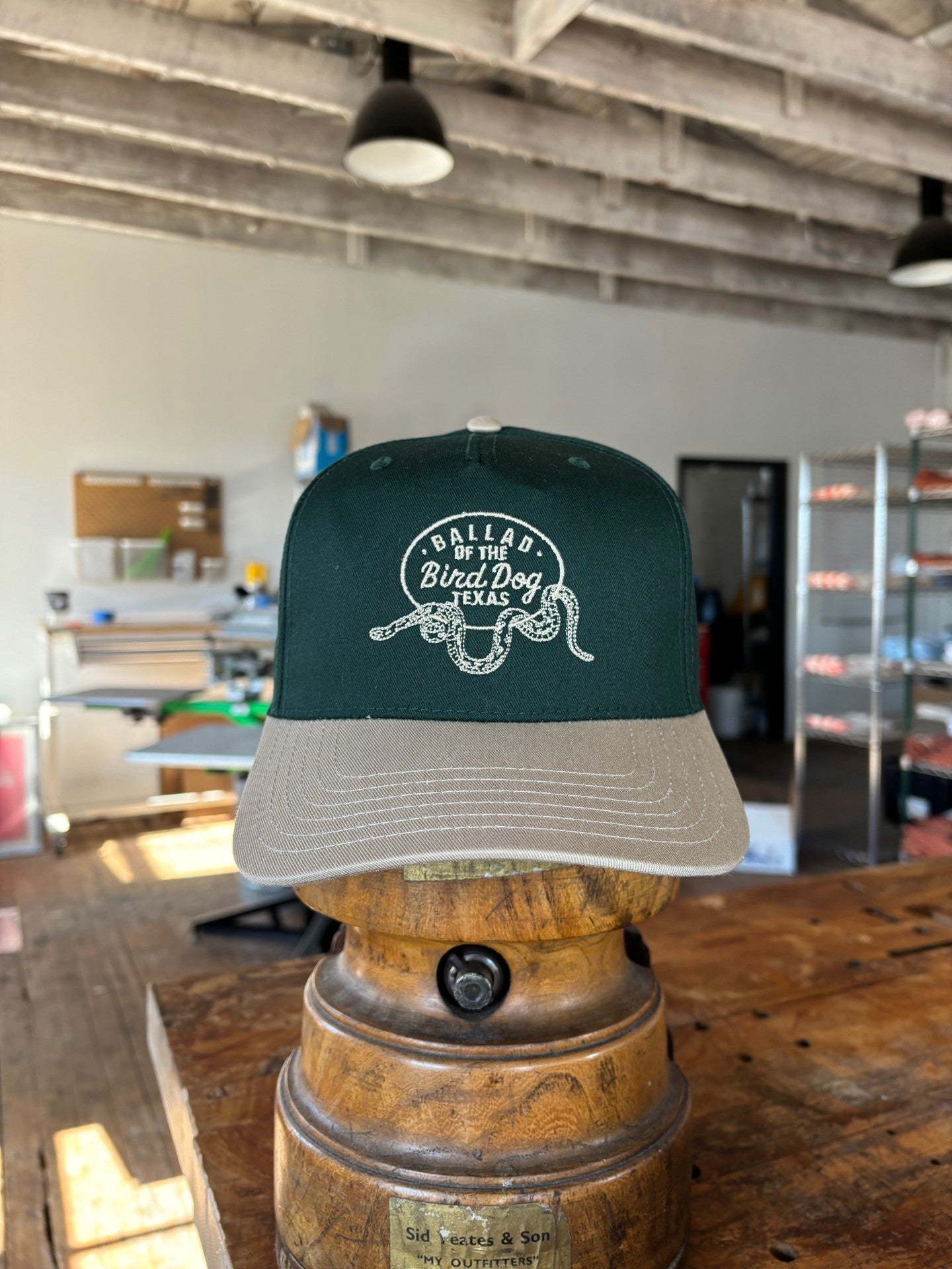 The Shop Hat | Rattlesnake Oval Logo by Ballad of The Bird Dog from the Desert Collection, in green and beige with a rattlesnake graphic and "Dallas by the Bird Dog, Texas," rests on a wooden stand in a workshop.