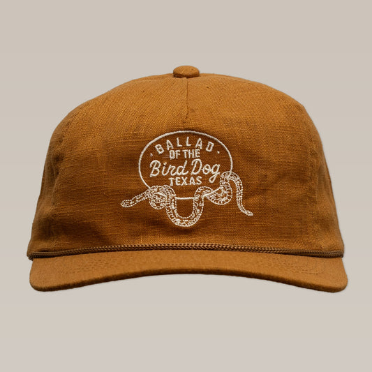 The Shop Hat by Ballad of the Bird Dog features a brown cap handcrafted in Brenham, TX, with a white "Ballad of the Bird Dog Texas" embroidery and bold rattlesnake oval logo—a perfect snake-themed hat for collectors.