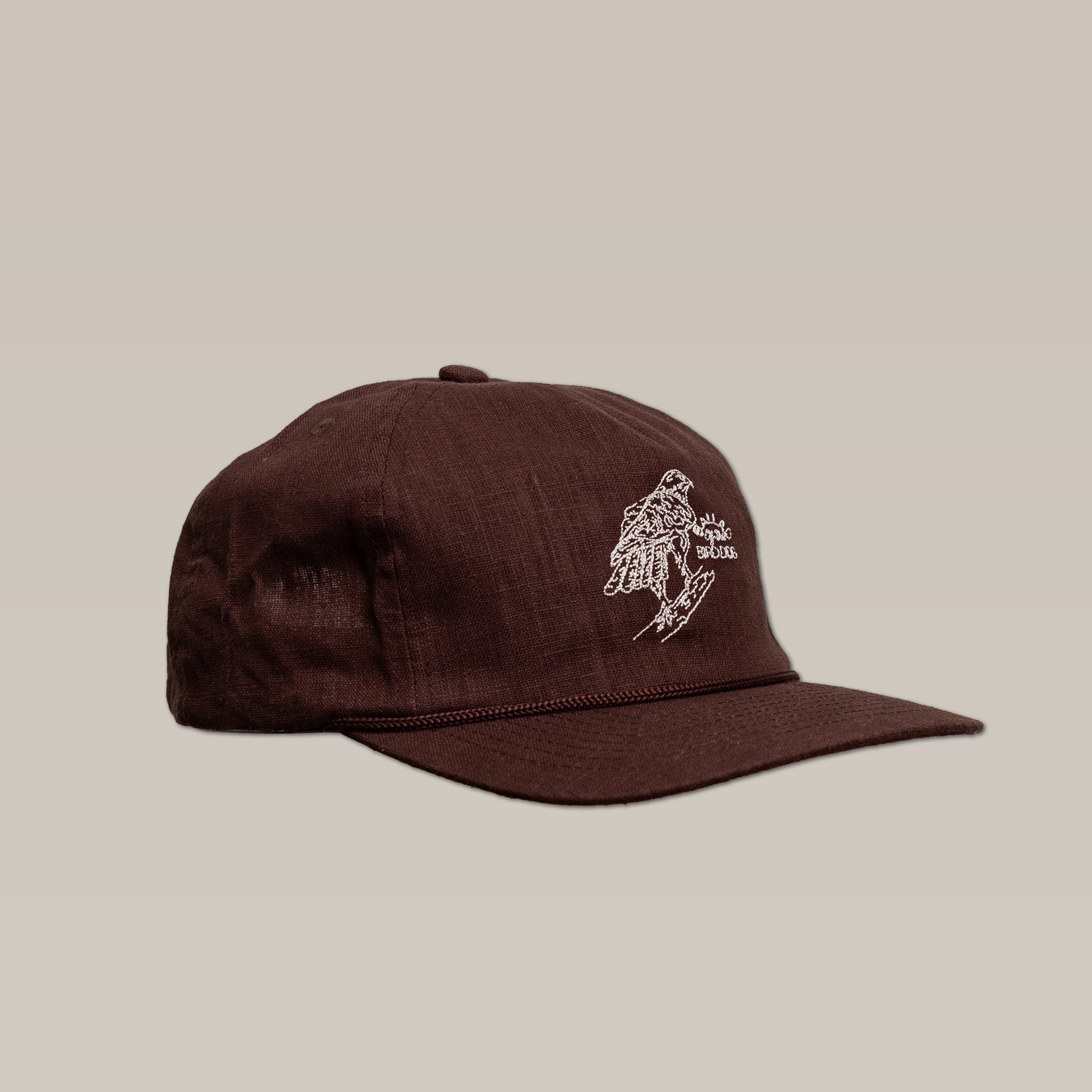 The Shop Hat | Red Tailed Hawk by Ballad of the Bird Dog is a brown baseball cap featuring white embroidered artwork of a hand and skateboard on the front, displayed against a plain beige background.