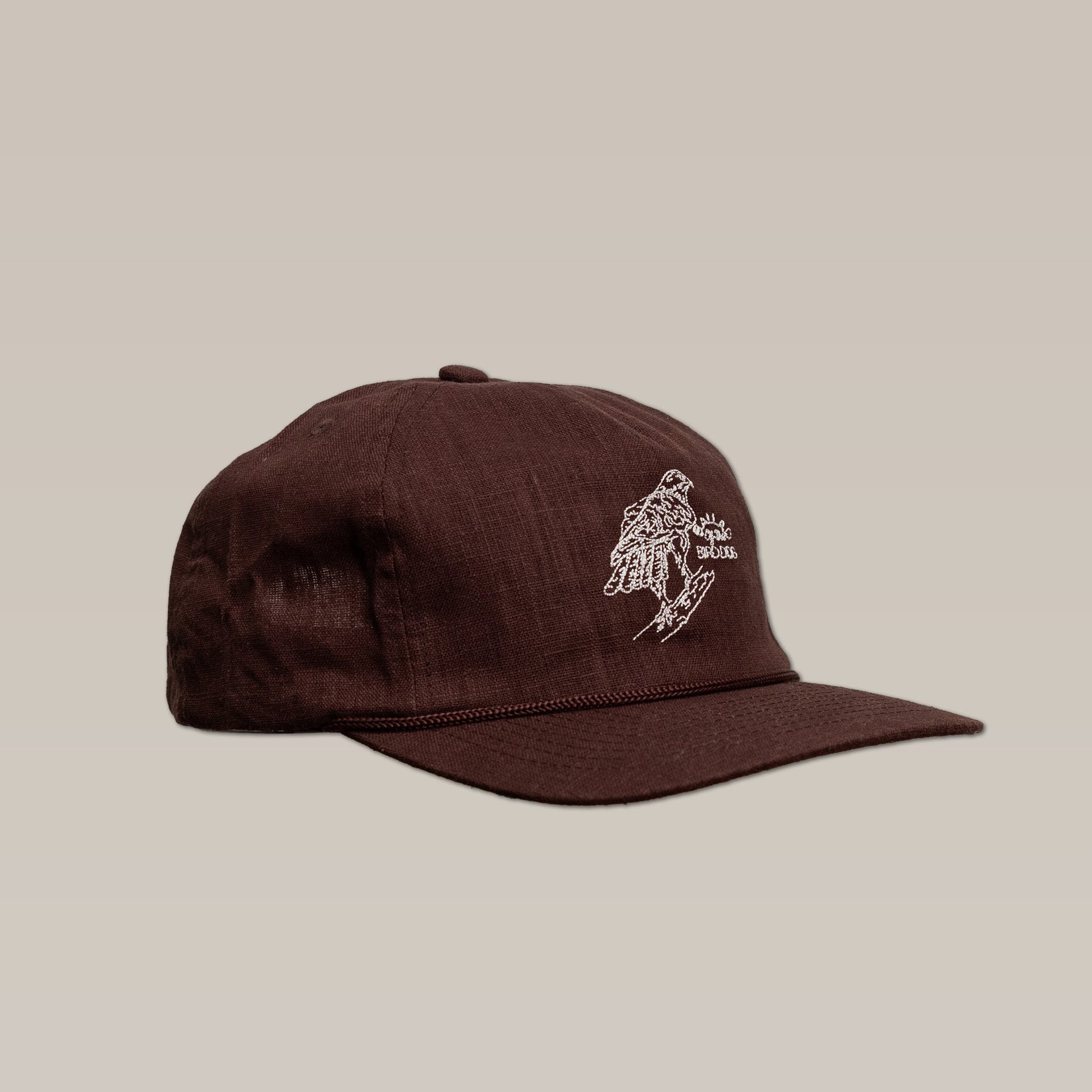 The Shop Hat | Red Tailed Hawk by Ballad of the Bird Dog is a brown baseball cap featuring a white embroidered hand design and text on the front, shown against a plain beige background.
