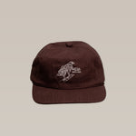 The Shop Hat | Red Tailed Hawk by Ballad of the Bird Dog is a brown baseball cap featuring a white embroidered bird design on the front, shown against a plain beige background.