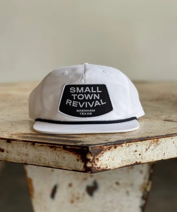 The Ballad of the Bird Dog Shop Hat features a "SMALL TOWN REVIVAL BRENHAM TEXAS" patch, making it perfect for small town patch hat fans. Shown here on a distressed wooden table.