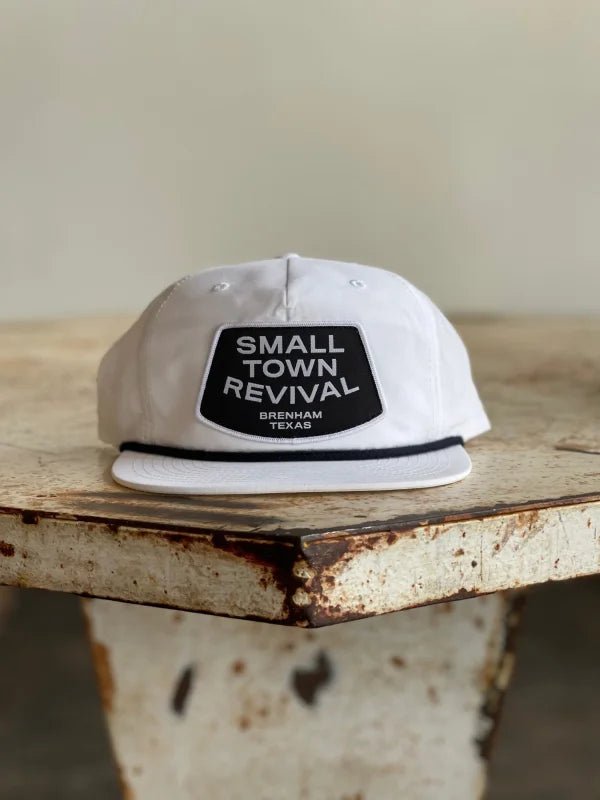 The Ballad of the Bird Dog Shop Hat features a "SMALL TOWN REVIVAL BRENHAM TEXAS" patch, making it perfect for small town patch hat fans. Shown here on a distressed wooden table.