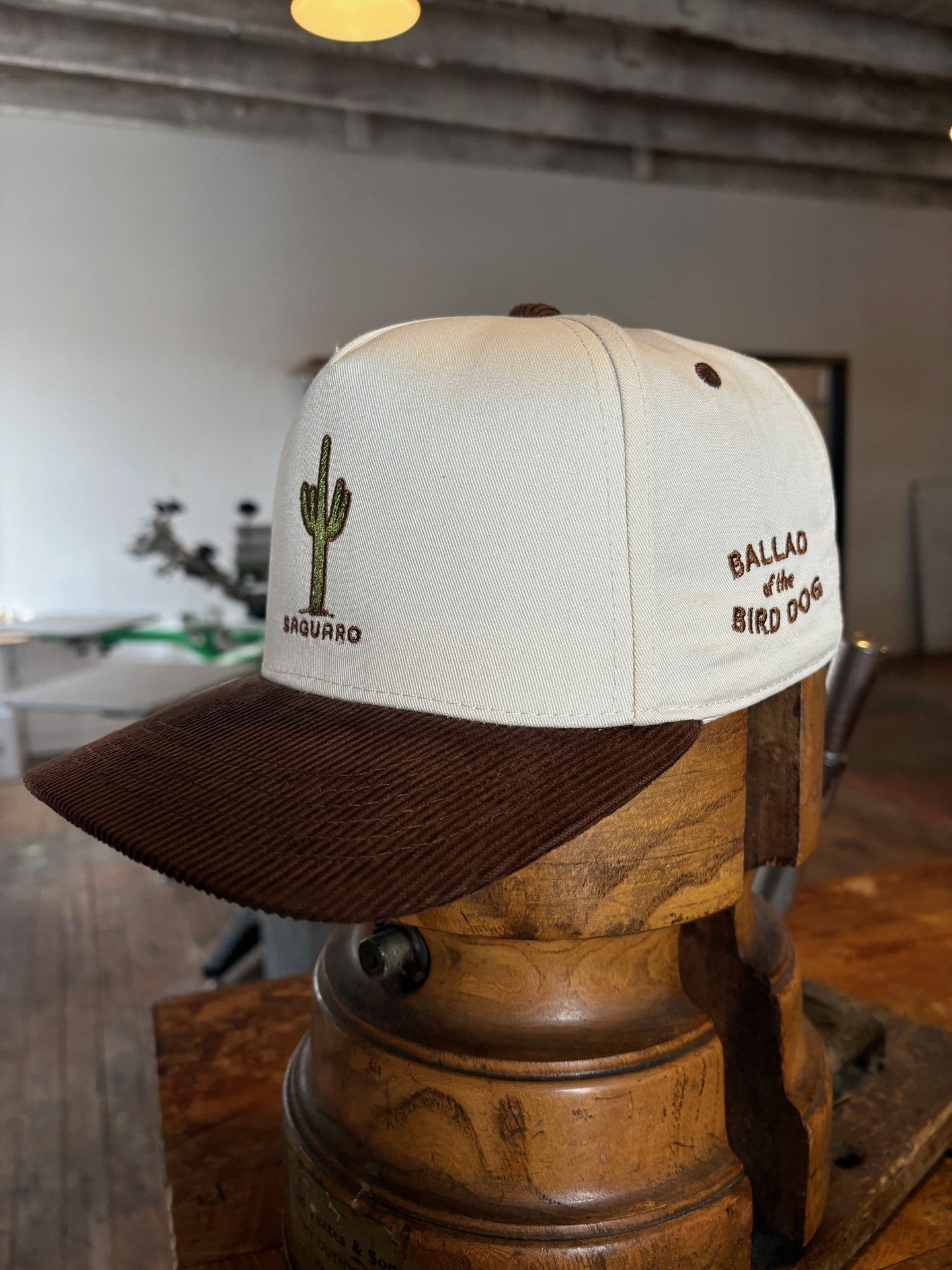 The Shop Hat | Saguaro by Ballad of the Bird Dog is a white and brown baseball cap with a cactus and "Saguaro" on the front, and "Ballad of the Bird Dog" embroidered on the side, shown indoors on a wooden hat stand.