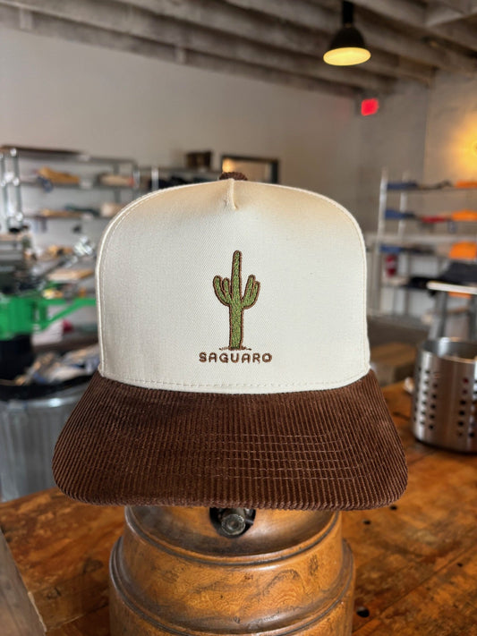 The Shop Hat | Saguaro by Ballad of the Bird Dog is a cream and brown trucker hat featuring a cactus and "Saguaro" embroidered on the front, shown indoors on a metal stand.