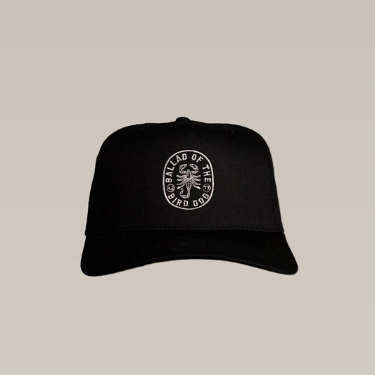 The Shop Hat | Scorpion by Ballad of the Bird Dog is a black cap from the Desert Collection, featuring a white embroidered circular "BALLAD OF THE BIRD DOG" logo with a central graphic, shown on a plain beige background.