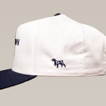 The Shop Hat by Ballad of the Bird Dog is a white baseball cap with a black brim, featuring a small embroidered dog on the side.