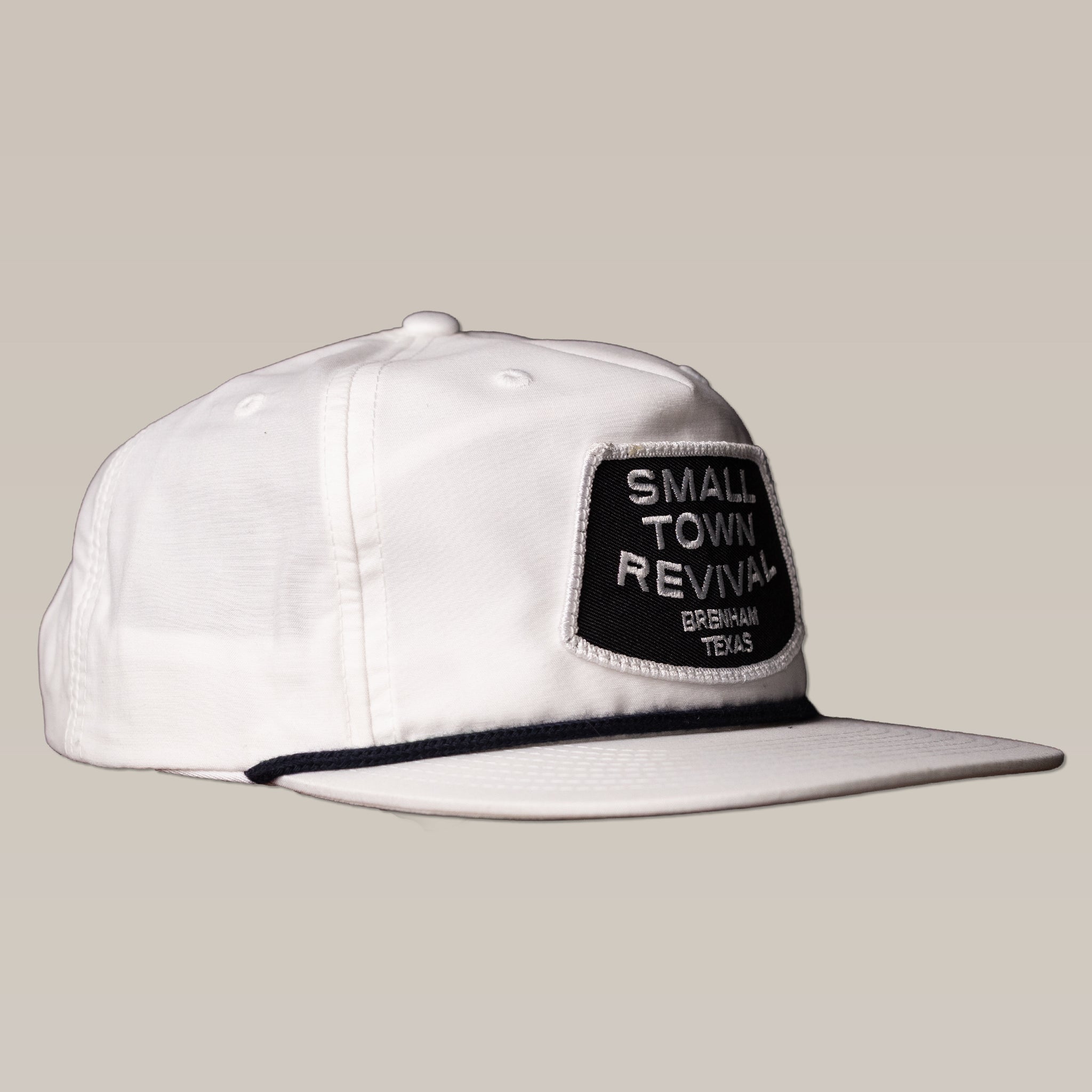 The Ballad of the Bird Dog Shop Hat features a black "Small Town Revival Brenham Texas" patch on a white snapback, perfect for small town patch hat fans. Shown on a plain beige background.