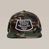 The Shop Hat by Ballad of the Bird Dog features a camouflage design with a white "SMALL TOWN REVIVAL BRENHAM TEXAS" patch—perfect for fans of small town pride and unique patch hats.