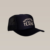 The Ballad of the Bird Dog Shop Hat | Somewhere Texas is a black mesh snapback trucker hat with "SOMEWHERE TEXAS" embroidered in white on the front, designed by Carra Sykes and shown against a beige background.