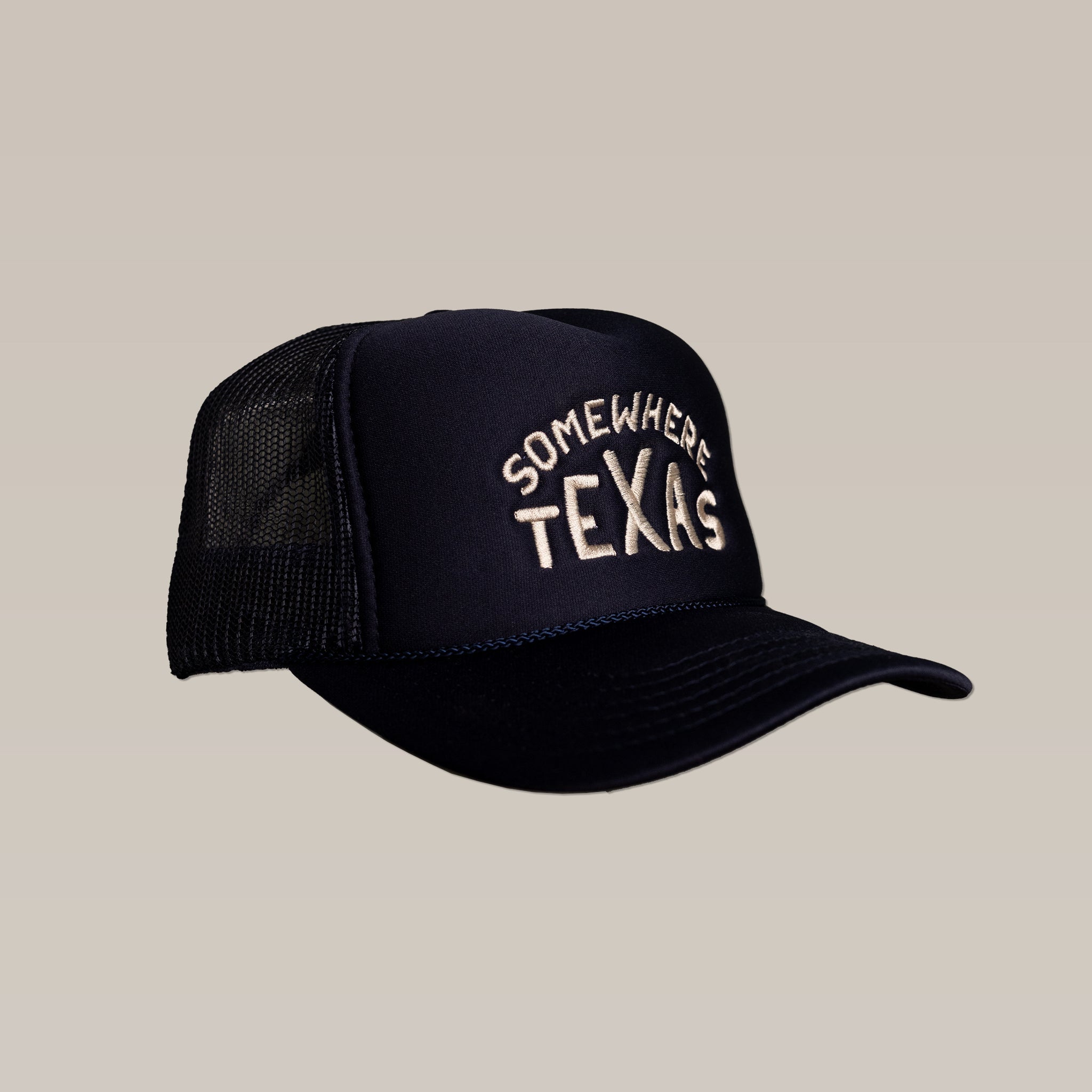 The Ballad of the Bird Dog Shop Hat | Somewhere Texas is a black mesh snapback trucker hat with "SOMEWHERE TEXAS" embroidered in white on the front, designed by Carra Sykes and shown against a beige background.