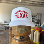 A white Shop Hat by Ballad of the Bird Dog, featuring bold red "Somewhere Texas" lettering, sits on a wooden hat block in a workshop with spools of thread and boxes in the background.