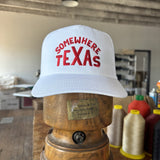 The Shop Hat | Somewhere Texas by Ballad of the Bird Dog features "SOMEWHERE TEXAS" in red on white. This classic snapback sits on a wooden block in a workshop, surrounded by spools and supplies—a must-have for fans.