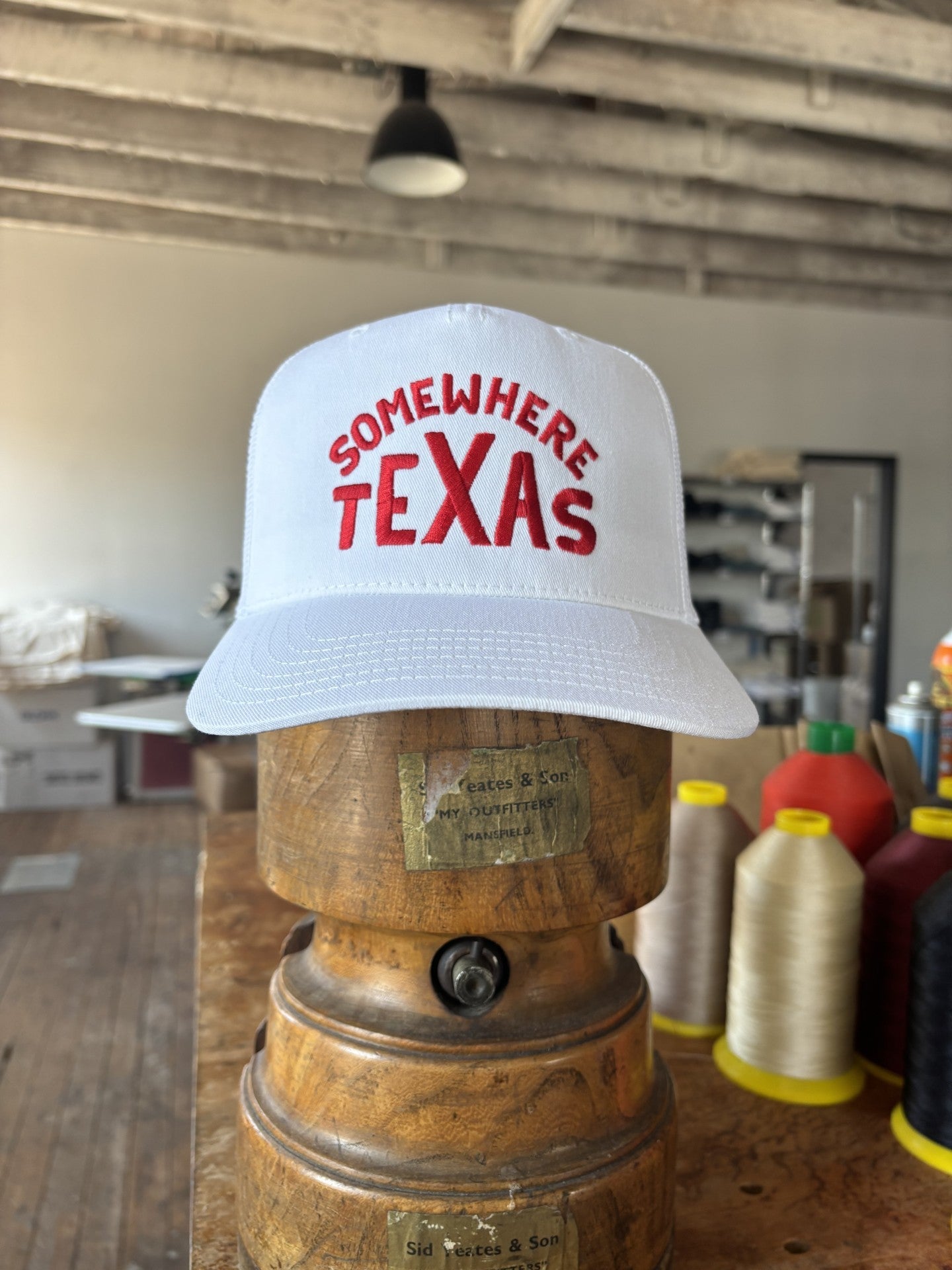 The Shop Hat | Somewhere Texas by Ballad of the Bird Dog features "SOMEWHERE TEXAS" in red on white. This classic snapback sits on a wooden block in a workshop, surrounded by spools and supplies—a must-have for fans.