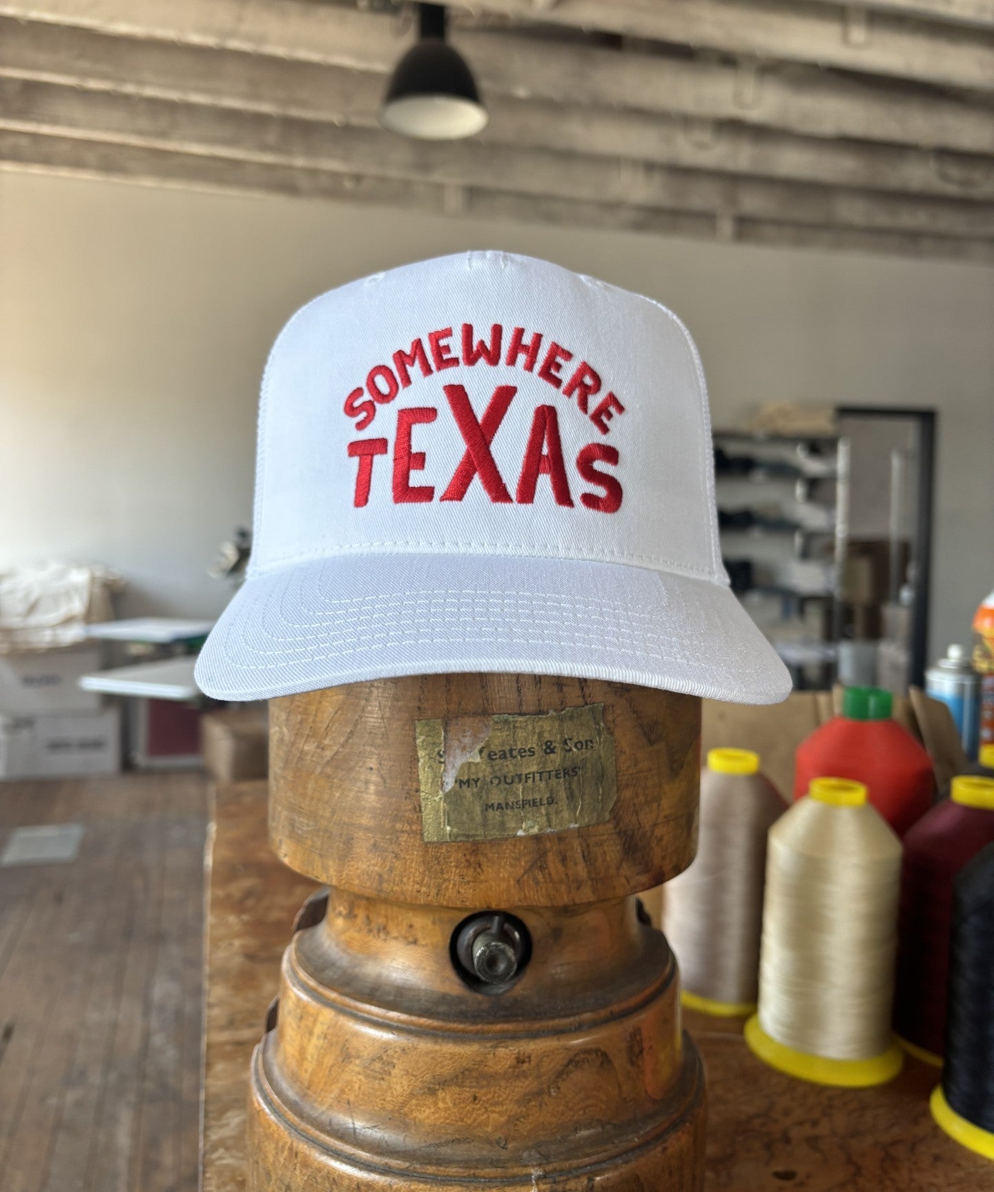A white Shop Hat | Somewhere Texas by Ballad of the Bird Dog, featuring “SOMEWHERE TEXAS” in red letters, rests on a wooden form in a workshop with threads and sewing supplies. Designed by Carra Sykes.