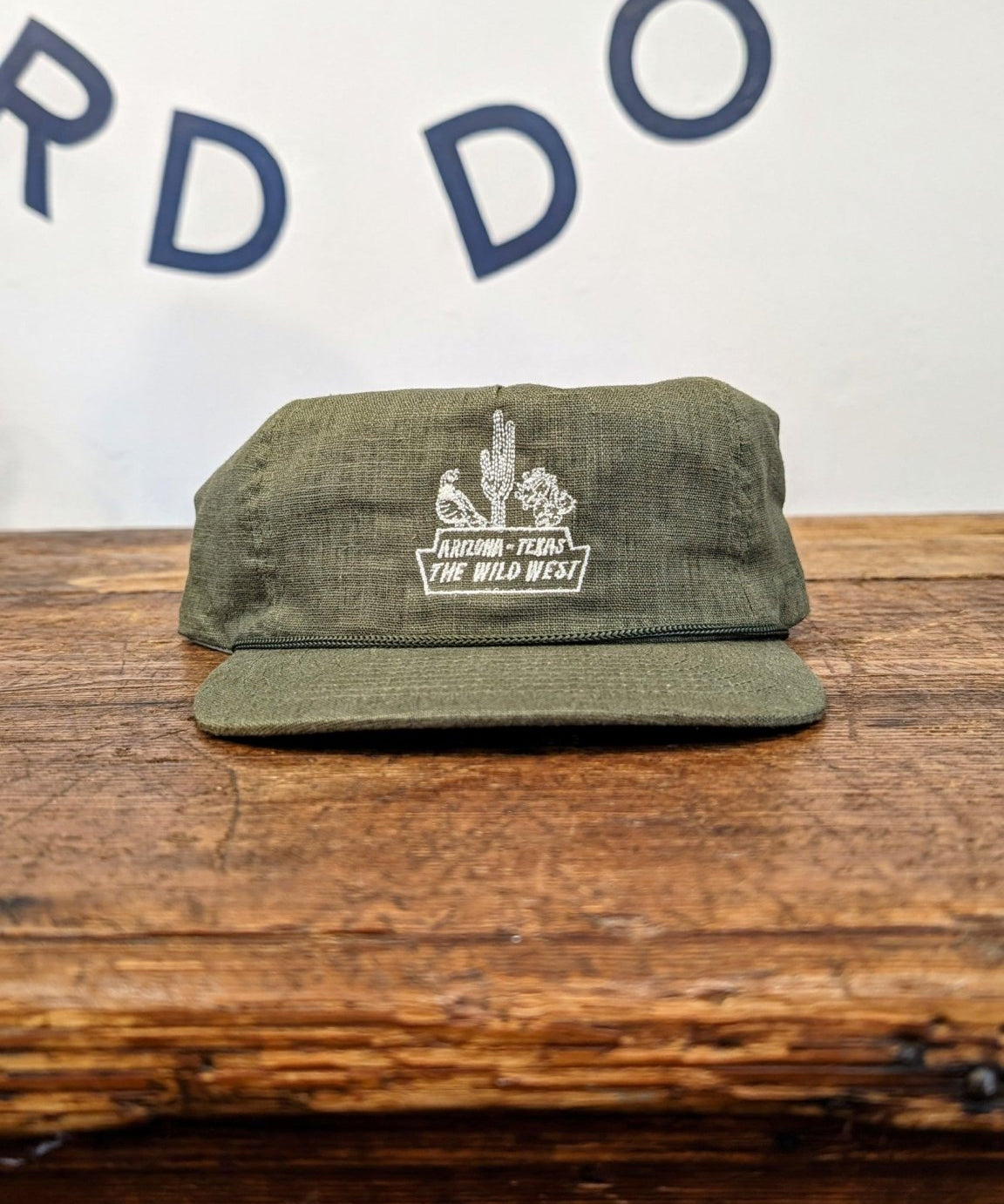 An olive green handcrafted Shop Hat from Ballad of the Bird Dog’s Desert Series Collection, featuring white embroidery with "ARIZONA, USA THE WILD WEST," rests on wood. Wall in background displays partial lettering.