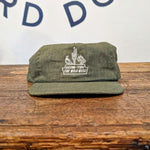 An olive green handcrafted Shop Hat from Ballad of the Bird Dog’s Desert Series Collection, featuring white embroidery with "ARIZONA, USA THE WILD WEST," rests on wood. Wall in background displays partial lettering.