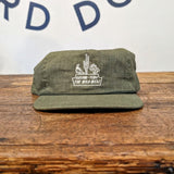An olive green handcrafted Shop Hat from Ballad of the Bird Dog’s Desert Series Collection, featuring white embroidery with "ARIZONA, USA THE WILD WEST," rests on wood. Wall in background displays partial lettering.