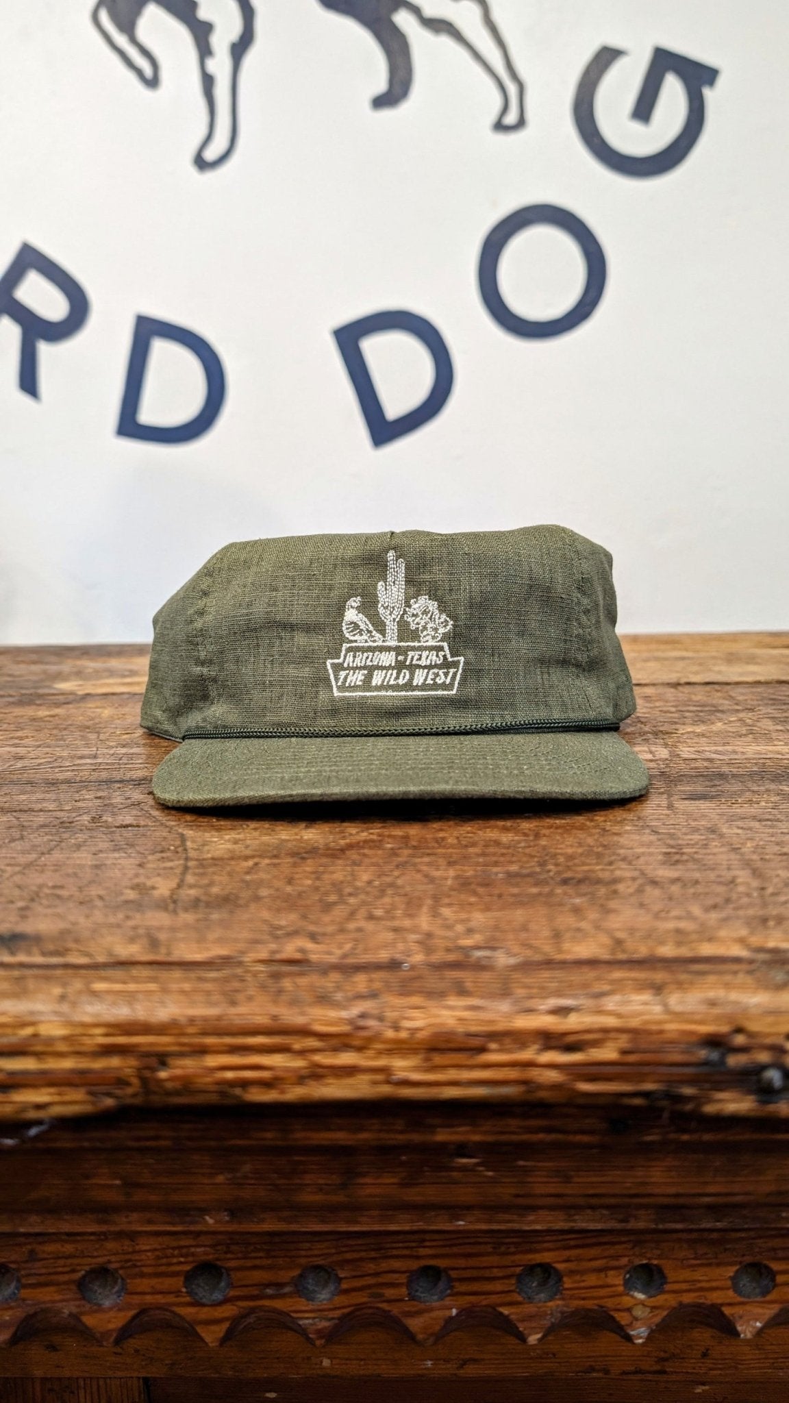 An olive green handcrafted Shop Hat from Ballad of the Bird Dog’s Desert Series Collection, featuring white embroidery with "ARIZONA, USA THE WILD WEST," rests on wood. Wall in background displays partial lettering.