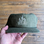 A hand holds the olive green Shop Hat | The Wild West by Ballad of the Bird Dog, with white "Arizona-Texas The Wild West" embroidery, cactus and horse details. Part of the Desert Series, it’s seen above wooden floorboards.