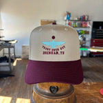 The Shop Hat | Thunderbird by Ballad of the Bird Dog, featuring beige and maroon colors, blue boat embroidery, and "TRUST YOUR GUT BRENHAM, TX" text, is displayed indoors on a metal stand.