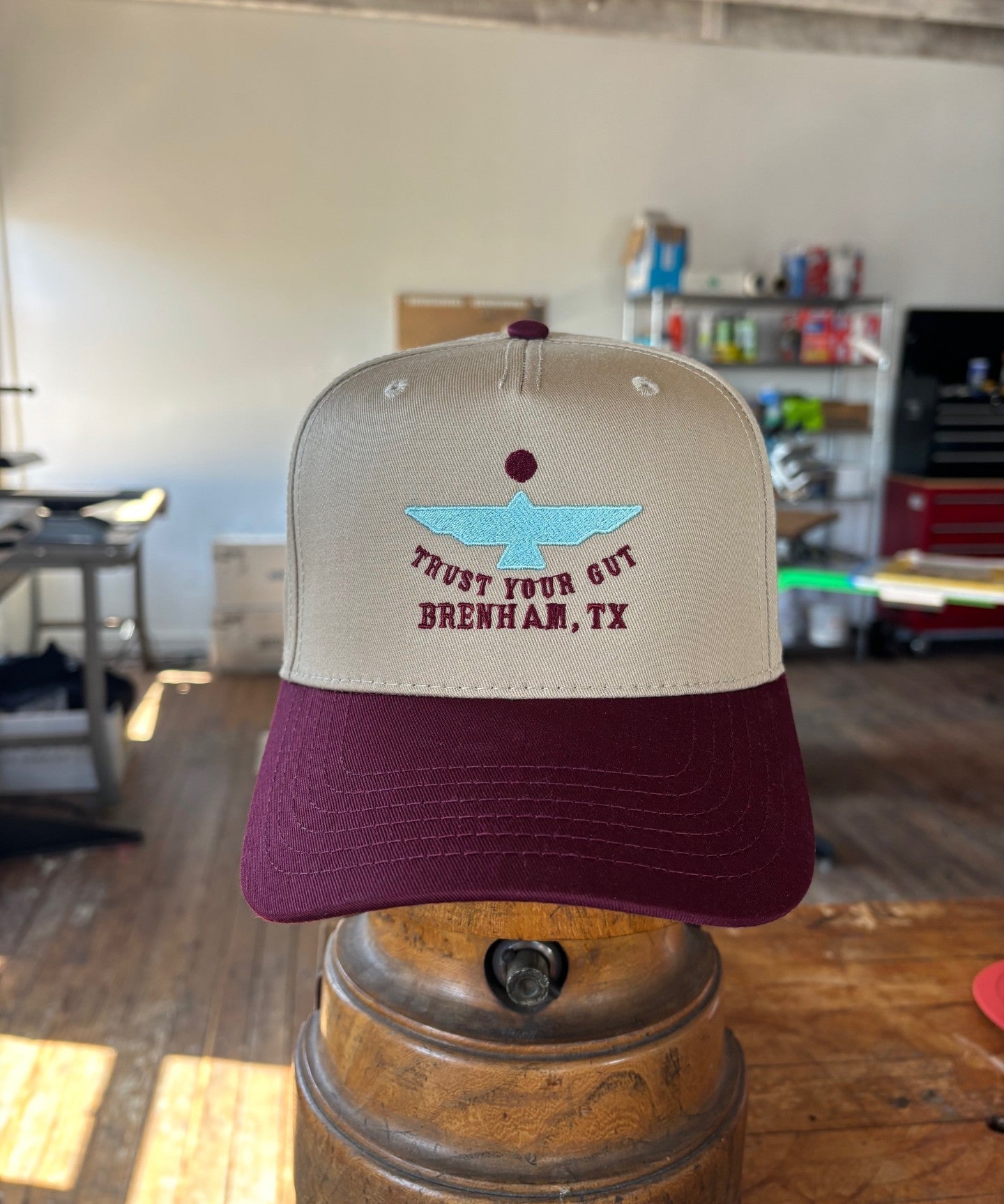 The Shop Hat | Thunderbird by Ballad of the Bird Dog, featuring beige and maroon colors, blue boat embroidery, and "TRUST YOUR GUT BRENHAM, TX" text, is displayed indoors on a metal stand.