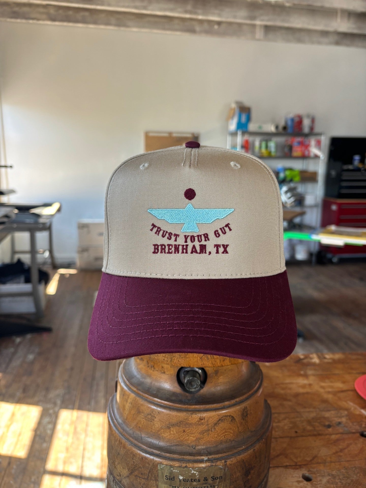 The Shop Hat | Thunderbird by Ballad of the Bird Dog, featuring beige and maroon colors, blue boat embroidery, and "TRUST YOUR GUT BRENHAM, TX" text, is displayed indoors on a metal stand.