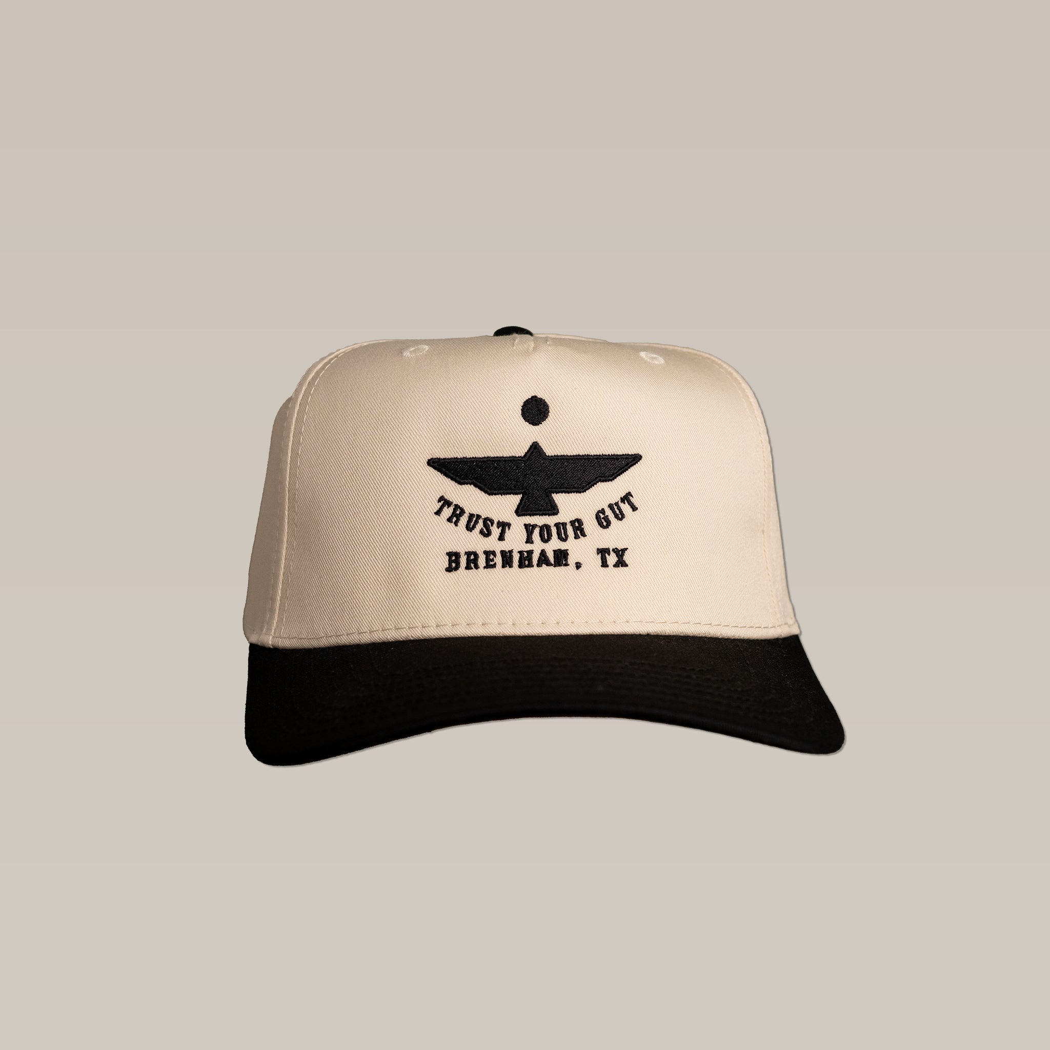 The Shop Hat | Thunderbird by Ballad of the Bird Dog is a beige and black cap with a bold thunderbird graphic and "TRUST YOUR GUT BRENHAM, TX" embroidered on the front.
