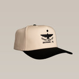 The Shop Hat | Thunderbird by Ballad of the Bird Dog is a beige and black cap with an abstract bird logo and "TRUST YOUR GUT BRENHAM, TX" embroidered on the front.