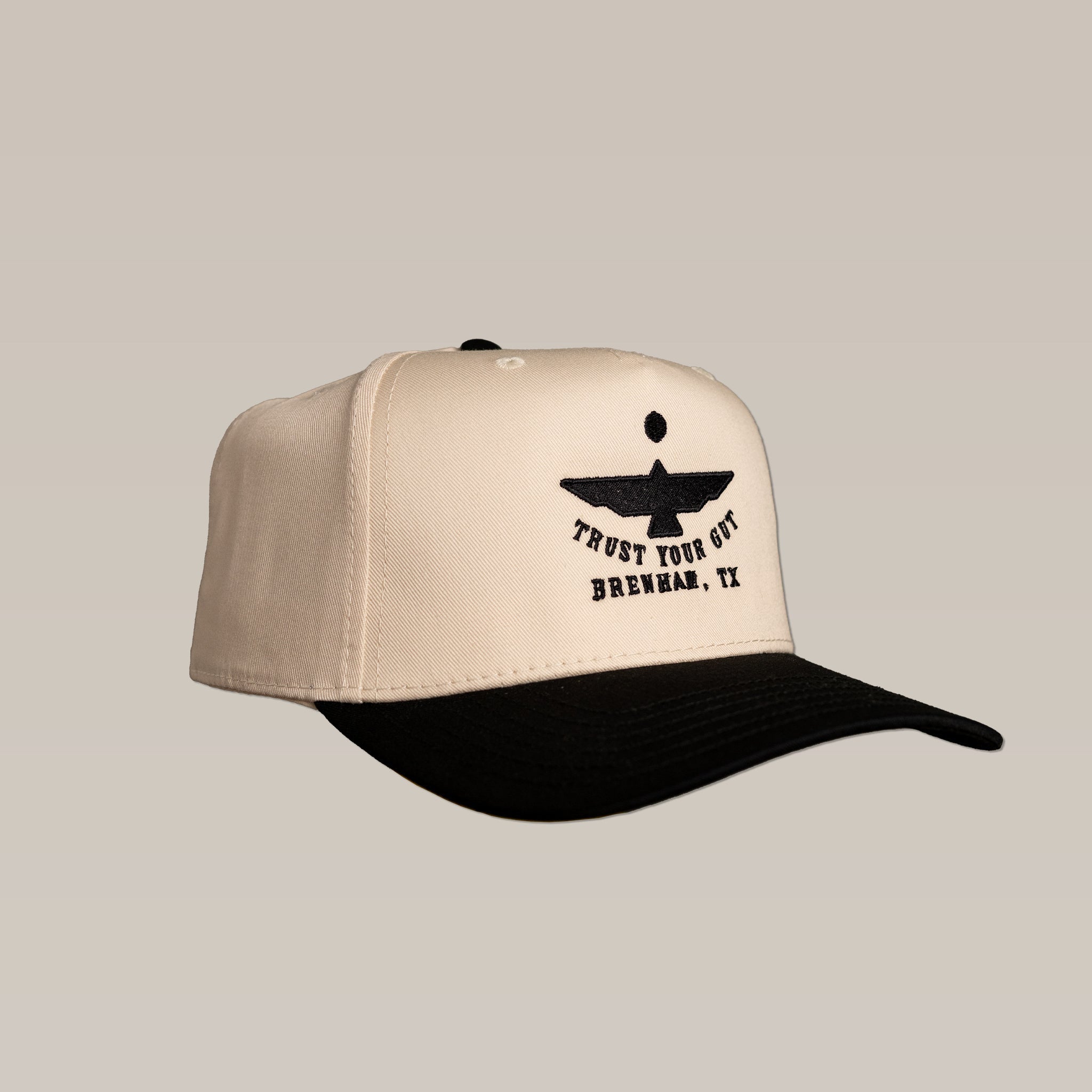 The Shop Hat | Thunderbird by Ballad of the Bird Dog is a beige and black cap with an abstract bird logo and "TRUST YOUR GUT BRENHAM, TX" embroidered on the front.