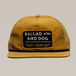 The Ballad of the Bird Dog Shop Hat | Trust Your Gut Vintage Slate is a mustard yellow snapback with a “BALLAD of the BIRD DOG” patch above “TRUST YOUR GUT,” inspired by the Trust Your Gut Vintage Slate Hat collection.