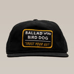 The Ballad of the Bird Dog Shop Hat in vintage slate is a black snapback featuring a rectangular patch with “BALLAD of the BIRD DOG 'TRUST YOUR GUT'” in white and yellow, inspired by their Trust Your Gut Vintage Slate design.