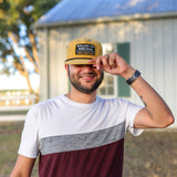 A man stands outside smiling, wearing a white and maroon shirt and a yellow cap, holding the brim of his Ballad of the Bird Dog Shop Hat | Trust Your Gut Vintage Slate. A building and trees are in the background.