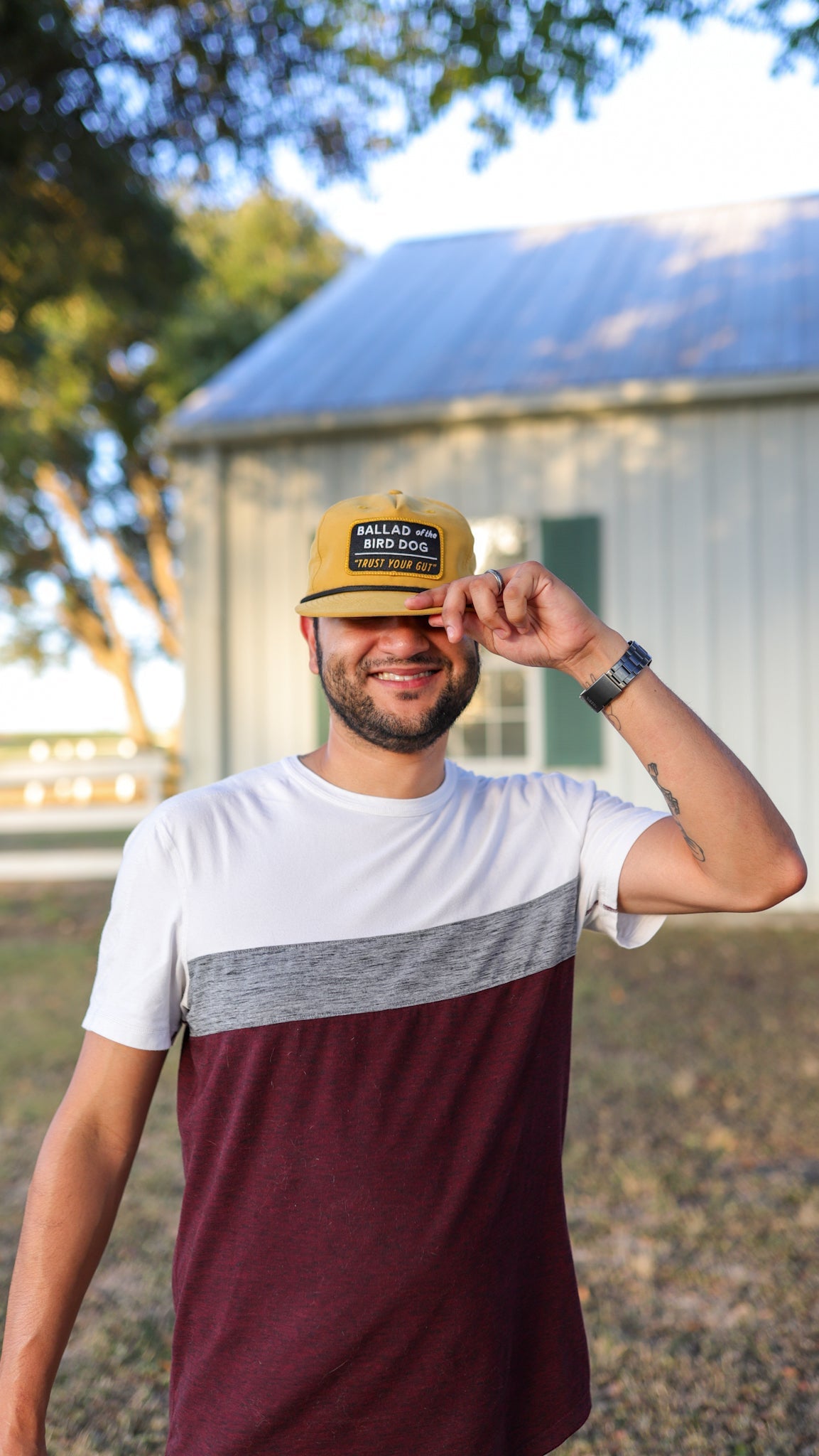 A man stands outside smiling, wearing a white and maroon shirt and a yellow cap, holding the brim of his Ballad of the Bird Dog Shop Hat | Trust Your Gut Vintage Slate. A building and trees are in the background.
