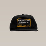 The Ballad of the Bird Dog Shop Hat in Trust Your Gut Vintage Slate features a yellow-bordered patch reading “BALLAD of the BIRD DOG ‘TRUST YOUR GUT’” on the front, shown on a plain beige background.