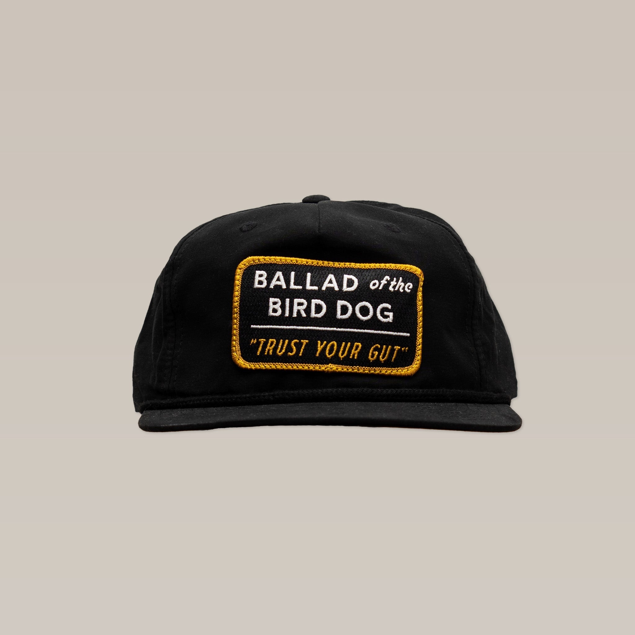 The Ballad of the Bird Dog Shop Hat in Trust Your Gut Vintage Slate features a yellow-bordered patch reading “BALLAD of the BIRD DOG ‘TRUST YOUR GUT’” on the front, shown on a plain beige background.