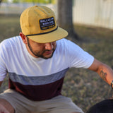 A man in a Trust Your Gut Vintage Slate Shop Hat by Ballad of the Bird Dog crouches outdoors in casual clothes, holding a red plastic cup, with part of a dog visible in the foreground.