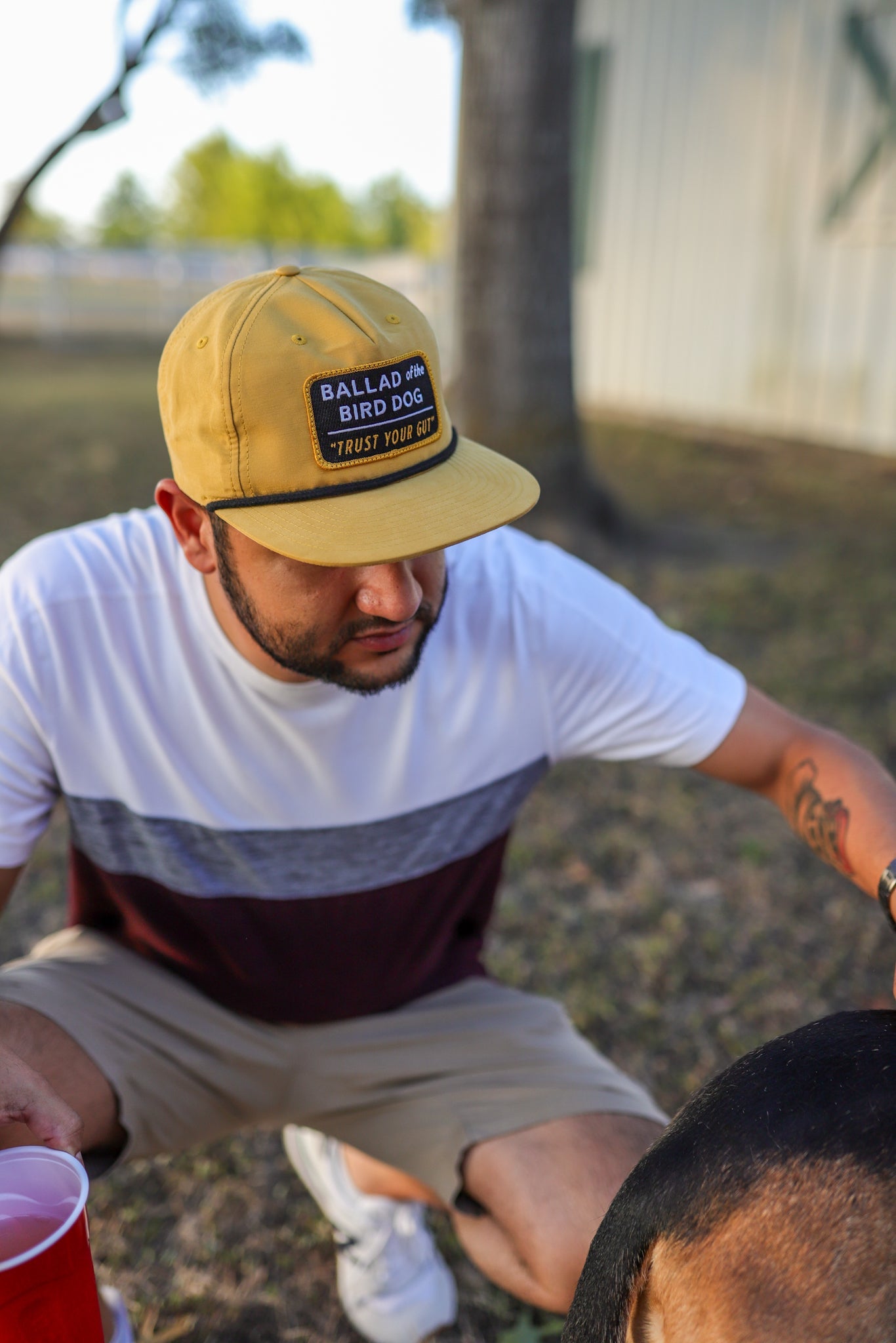 A man in a Trust Your Gut Vintage Slate Shop Hat by Ballad of the Bird Dog crouches outdoors in casual clothes, holding a red plastic cup, with part of a dog visible in the foreground.