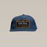 The Shop Hat | Trust Your Gut Vintage Slate from Ballad of the Bird Dog is a blue snapback featuring a rectangular patch with “BALLAD of the BIRD DOG ‘TRUST YOUR GUT’” in white and yellow—perfect for your collection.