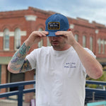 A man in a white Ballad of the Bird Dog tee and blue cap stands outside, holding the brim of his Ballad of the Bird Dog Shop Hat | Trust Your Gut Vintage Slate hat, with brick buildings in the background.