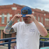 A man with a tattooed arm adjusts his Ballad of the Bird Dog Shop Hat | Trust Your Gut Vintage Slate and wears a white Ballad of the Bird Dog t-shirt, standing outdoors with brick buildings behind him.