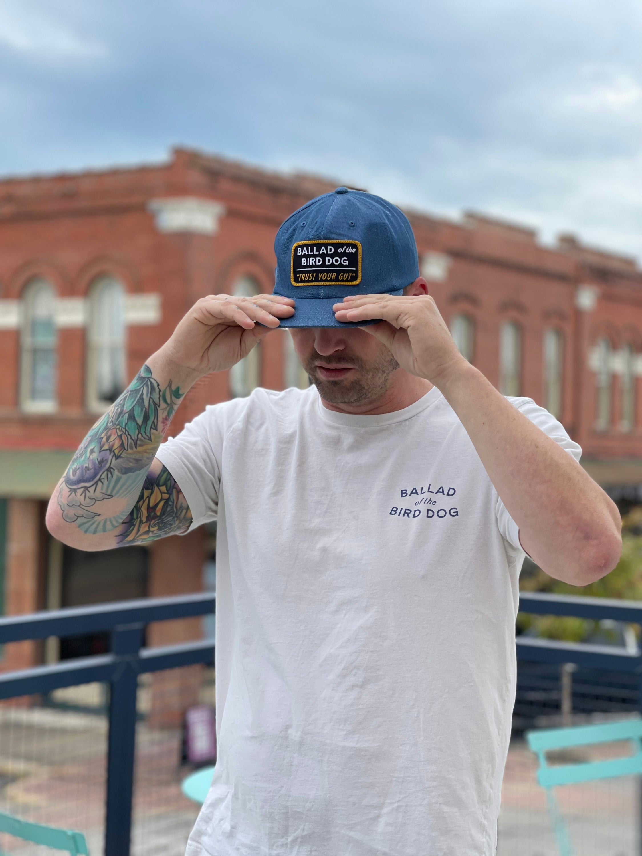 A man with a tattooed arm adjusts his Ballad of the Bird Dog Shop Hat | Trust Your Gut Vintage Slate and wears a white Ballad of the Bird Dog t-shirt, standing outdoors with brick buildings behind him.