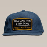 The Shop Hat | Trust Your Gut Vintage Slate by Ballad of the Bird Dog features a blue design and a rectangular patch reading “Ballad of the Bird Dog ‘Trust Your Gut’” in white and yellow letters.