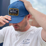 A man in a white t-shirt and the Ballad of the Bird Dog Shop Hat | Trust Your Gut Vintage Slate stands outdoors, adjusting his snapback hat. Both items feature the Ballad of the Bird Dog branding.