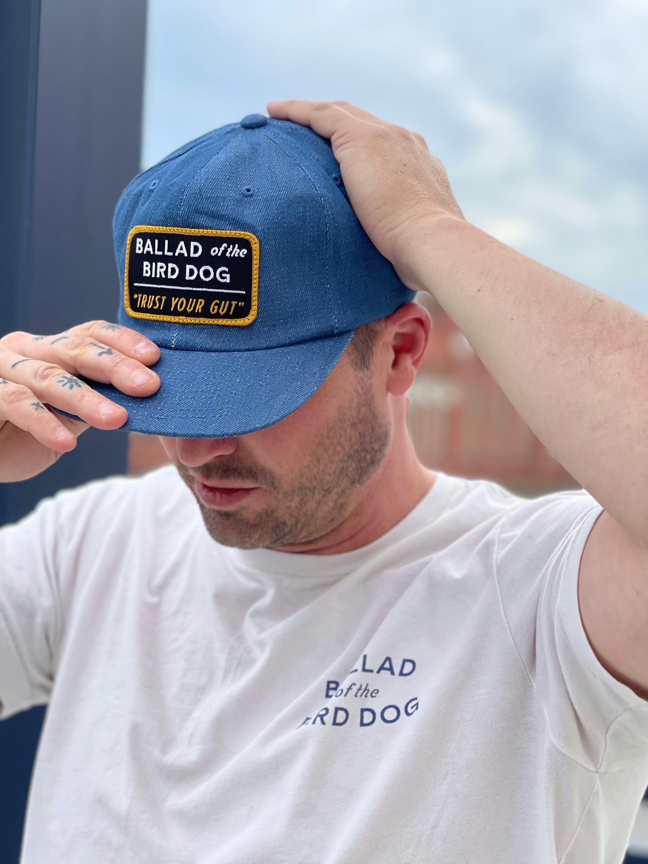 A man outdoors adjusts his blue "Shop Hat | Trust Your Gut Vintage Slate" cap and wears a white T-shirt, both branded "Ballad of the Bird Dog.